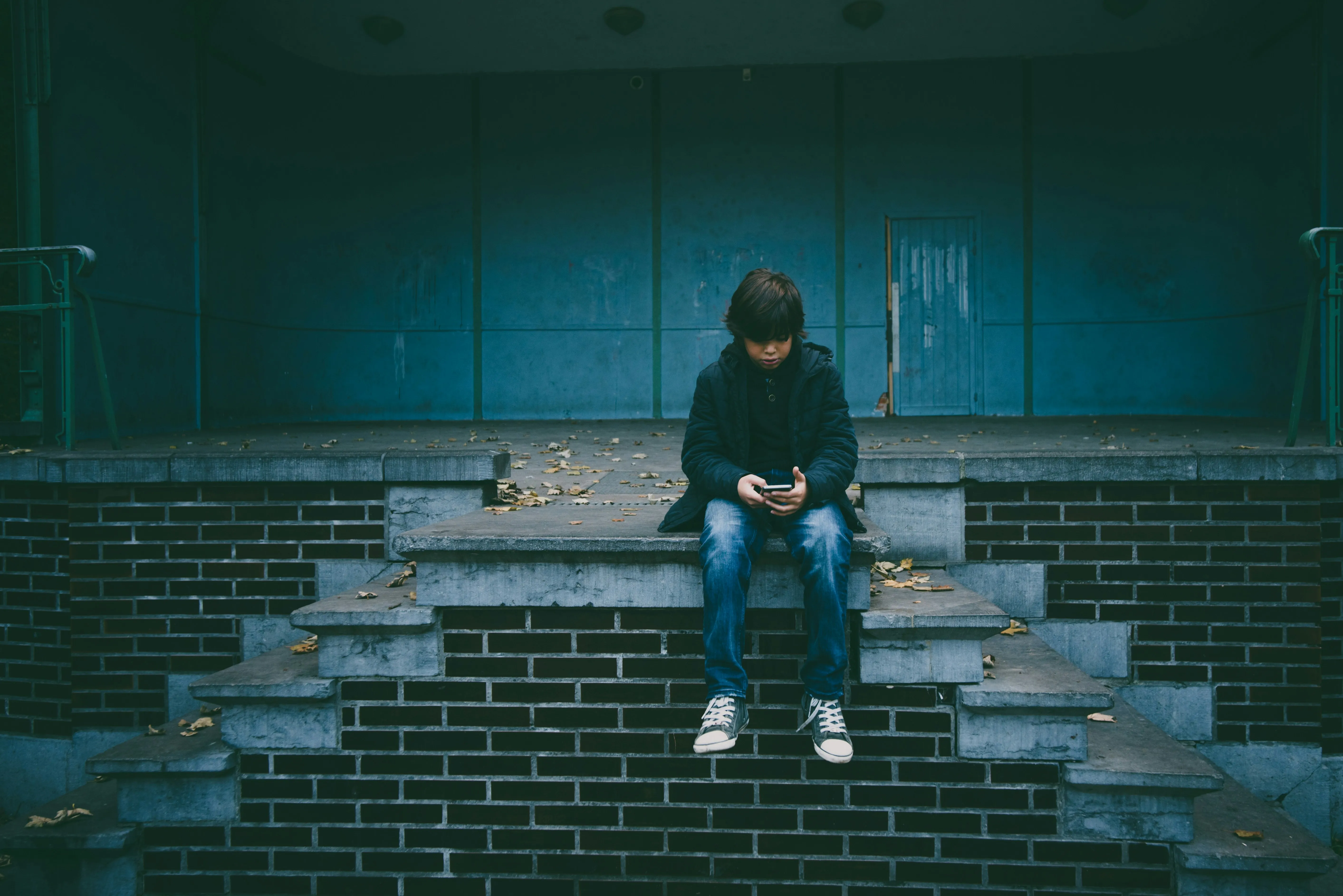A child sits alone on an outdoor staircase, with a mobile phone in their hands