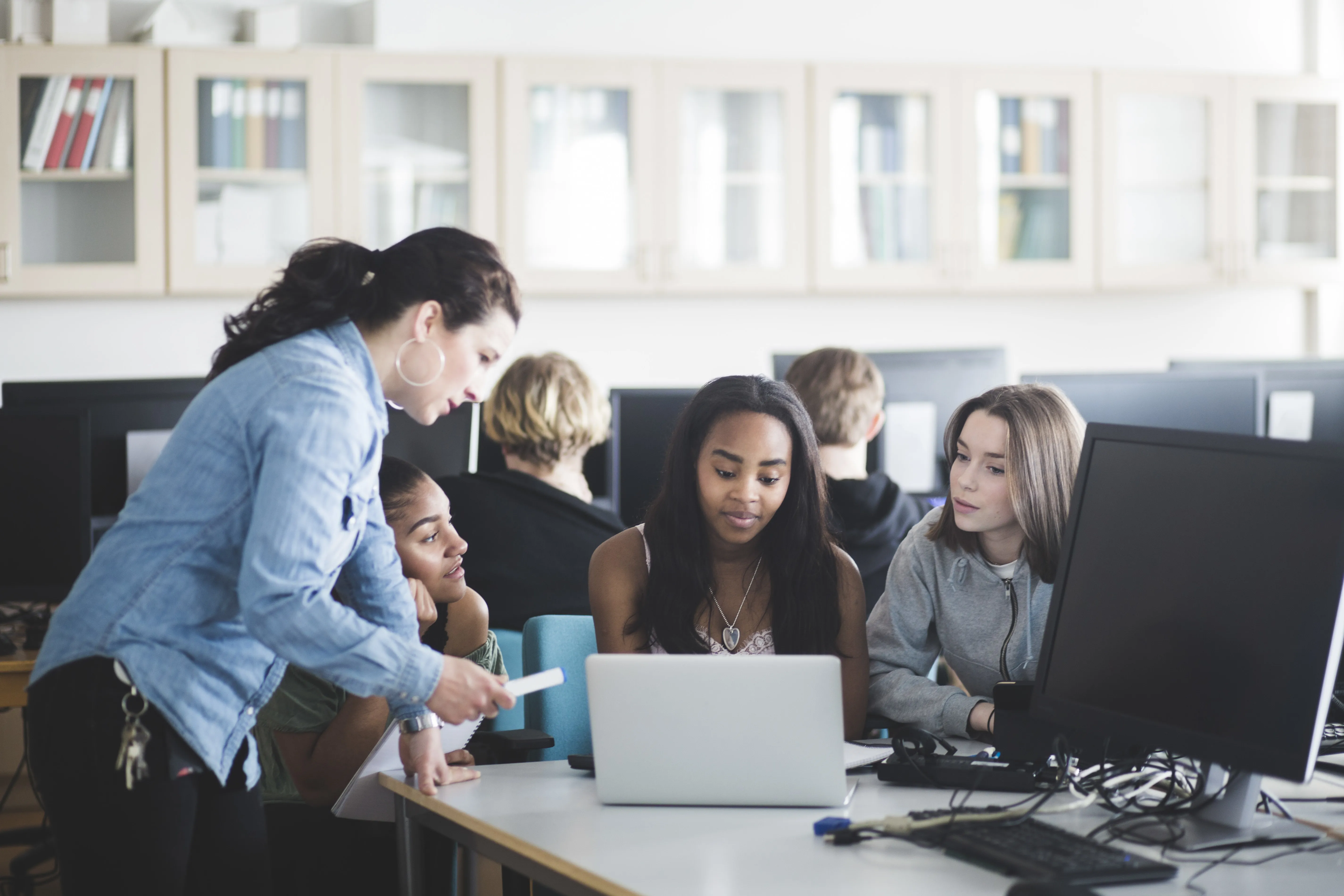 A female school teacher in a computer lab speaking to three teenage girls sitting in front of a laptop. 