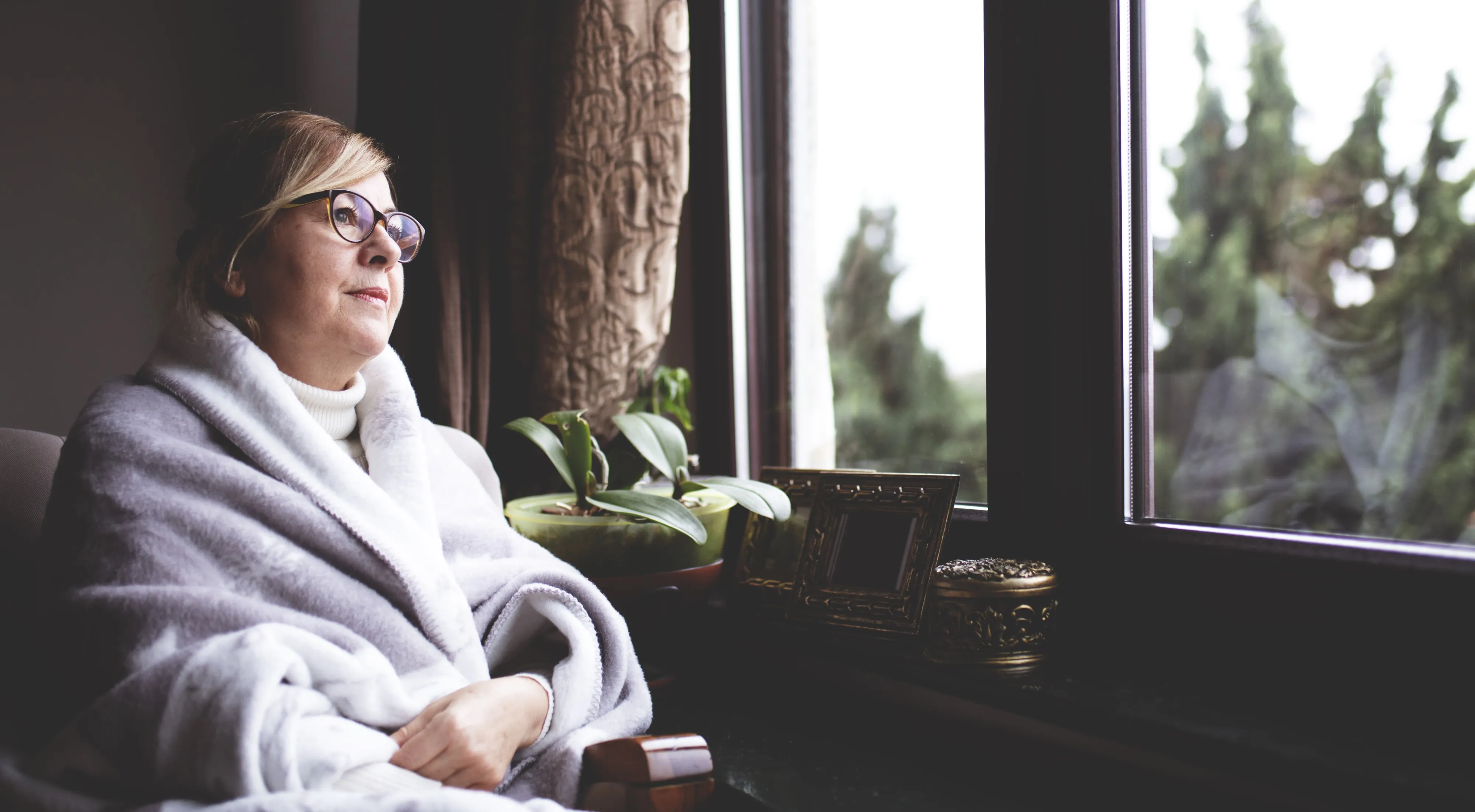 Woman sitting in a chair looks thoughtfully out of the window, wrapped in a blanket