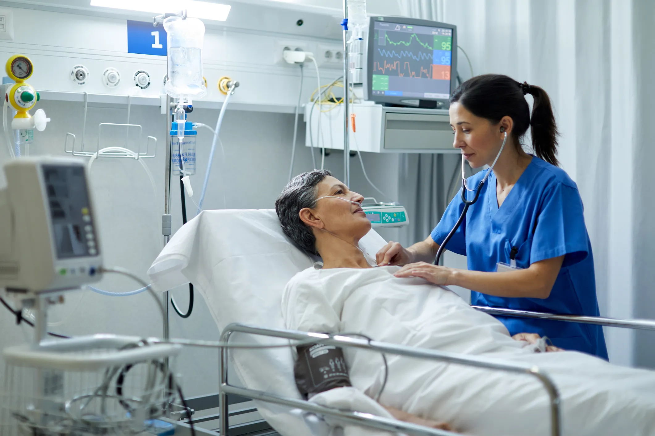 A female nurse is listening with a stethoscope to the heart of a patient in a hospital bed