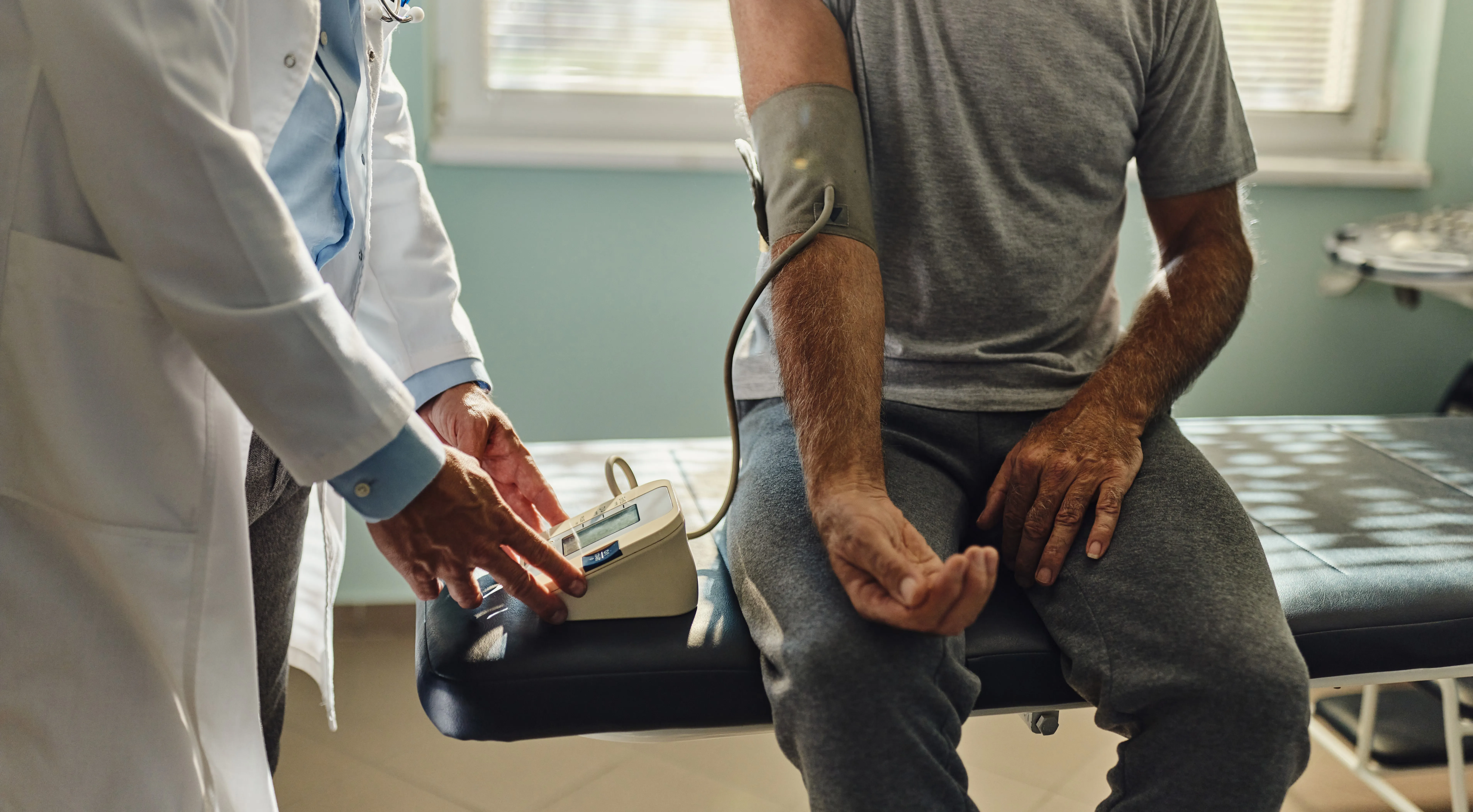 Male doctor in white lab coat taking blood pressure reading of male patient sat on hospital bed
