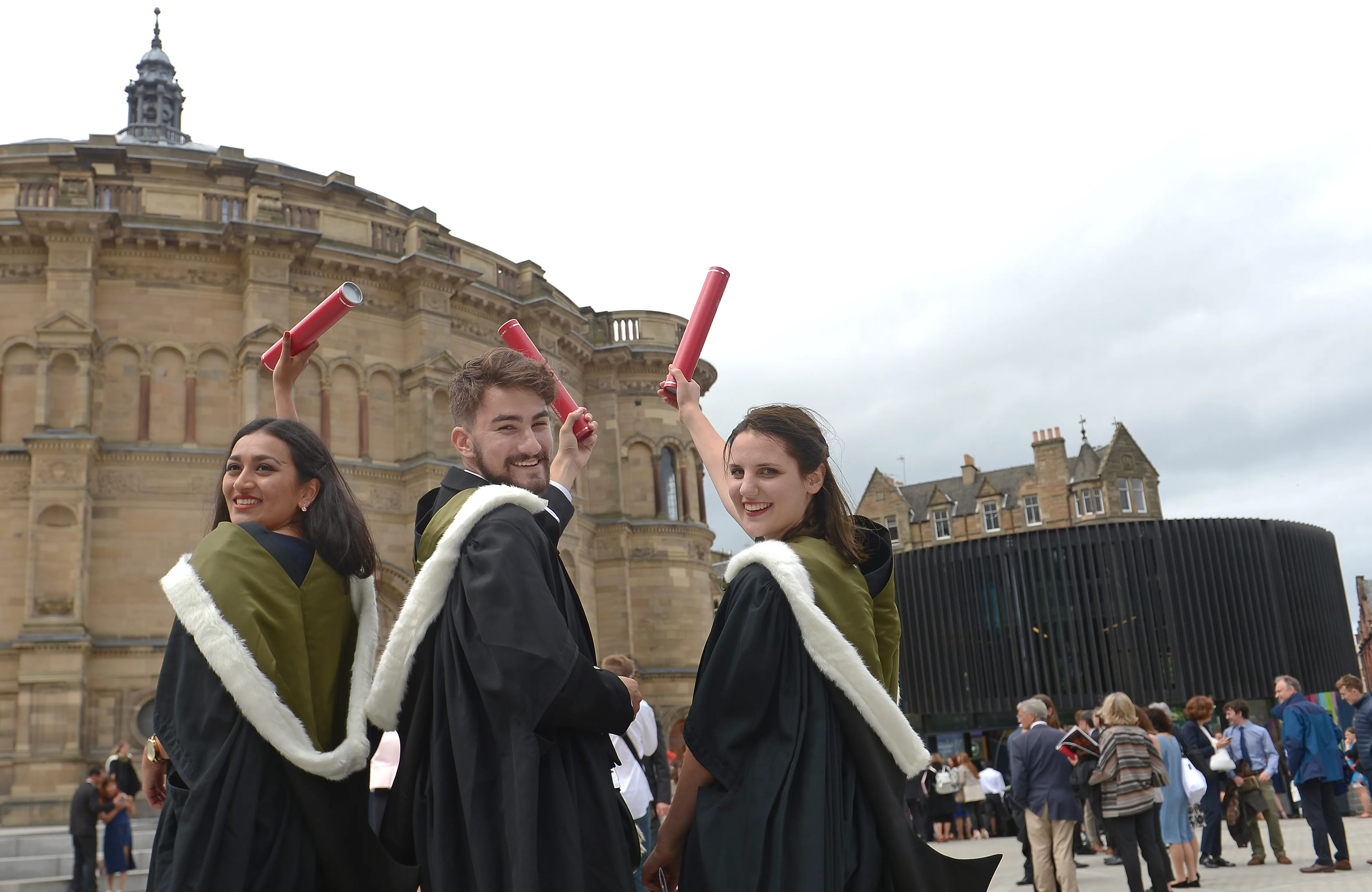 Three students celebrate with their families after graduation, holding red scrolls up to the sky.