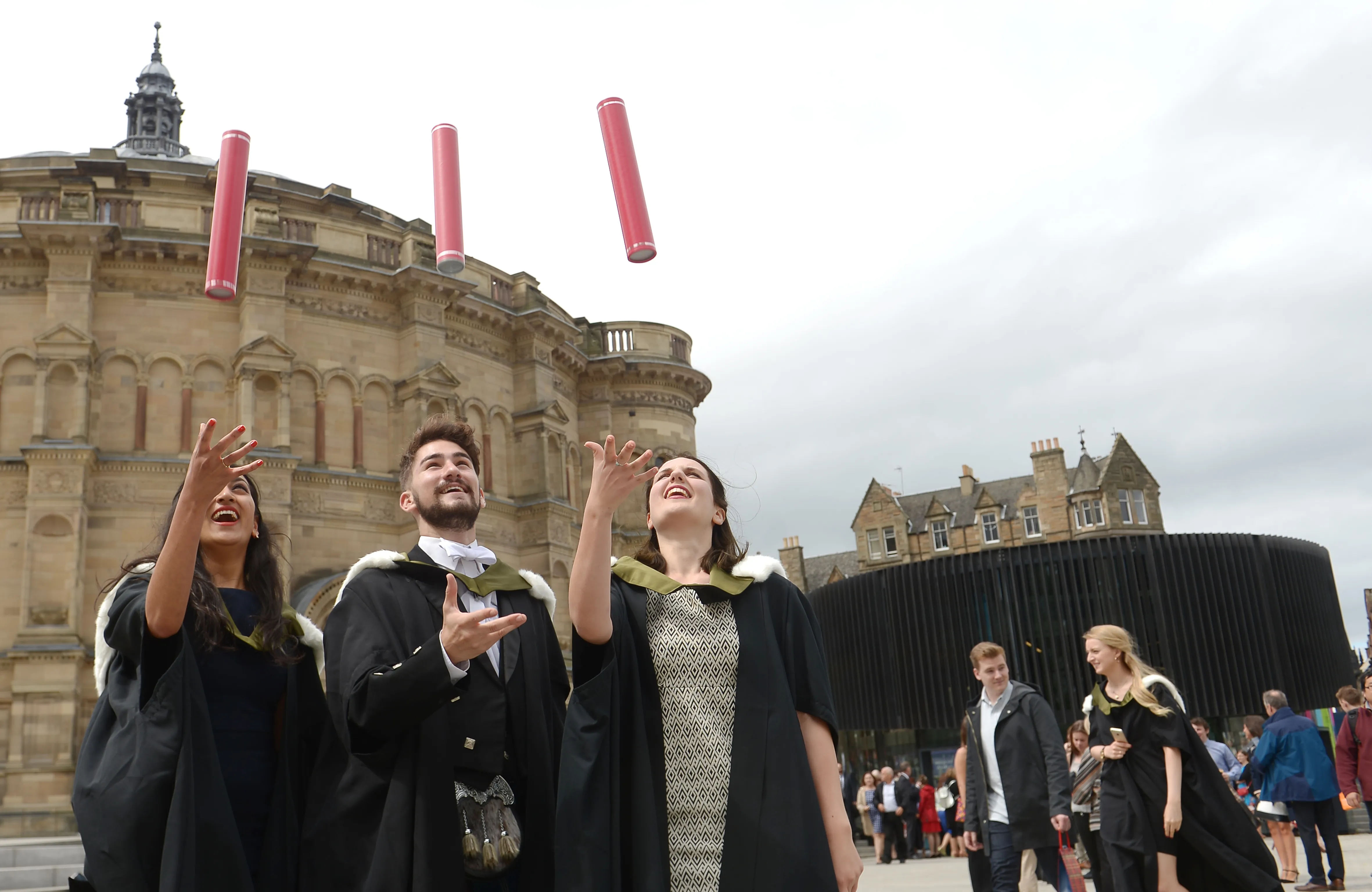 Three graduates standing together and celebrating their graduation by throwing red cylinder tubes in the air 