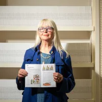 Cultural Activist Claudia Zeiske pictured holding a copy of her recipe book 'Combat Culture', in front of white shelving holding other copies.