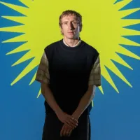Artist Eoghan Ryan pictured standing in front of a graphic yellow sun on a blue background, a still from his installation Terrible Simplifiers