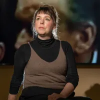 Filmmaker Jenny Brady pictured sitting in front of a blurred still featuring two people from her film The Glass Booth. 