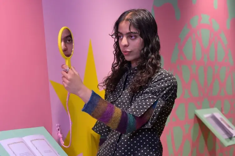A student artist poses in front of their installation looking in a mirror. 