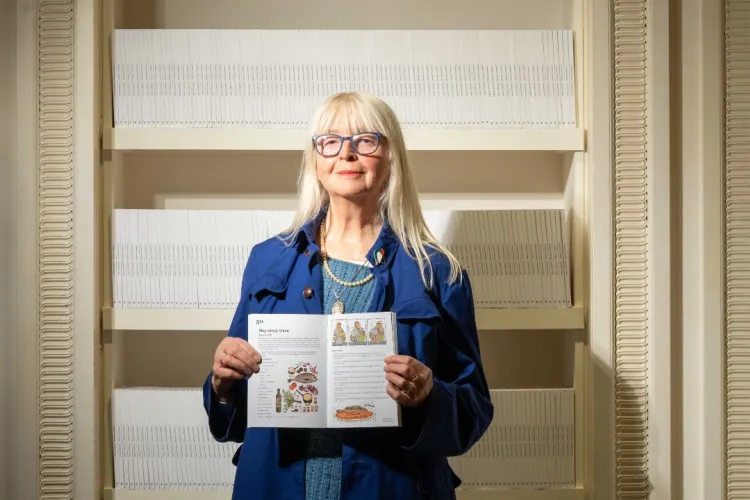 Cultural Activist Claudia Zeiske pictured holding a copy of her recipe book 'Combat Culture', in front of white shelving holding other copies.