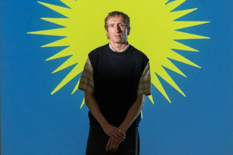 Artist Eoghan Ryan pictured standing in front of a graphic yellow sun on a blue background, a still from his installation Terrible Simplifiers