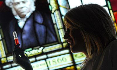 A blonde woman looks at a red test tube in front of a stained glass portrait of William Dick; the year 1866 is written below the portrait