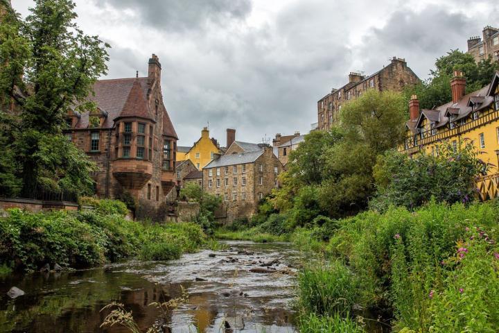 Photograph of Dean Village in Edinburgh. At the bottom of the photo is the Water of Leith with trees around the side. At the top of the picture are houses that look onto the Water of Leith. 