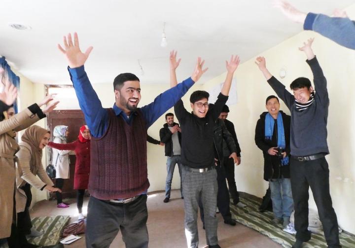 Photograph of a group of Afghan Peace Volunteers in a room doing a capacitar practice. All of the people in the group are raising their arms to the sky. 