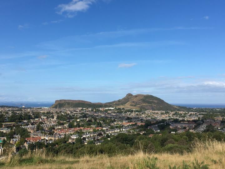 A photograph of Edinburgh with Arthur's Seat in the background. There are blue skies overhead and in the distance is the Firth of Forth 