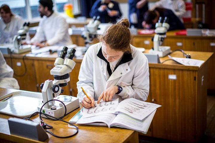 A student in a white lab coat writing in her lab book, with a microscope beside her.