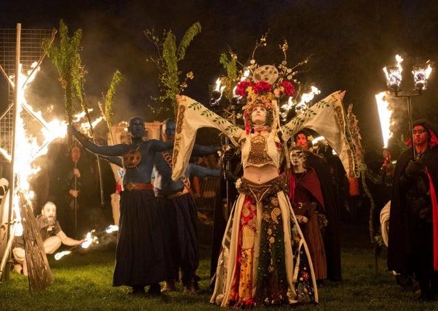 Photograph of May Queen at the Beltane Fire Festival. In the middle of the photograph is a woman in an elaborate costume surrounded by other Beltane performers, in the background are people holding lit torches. 