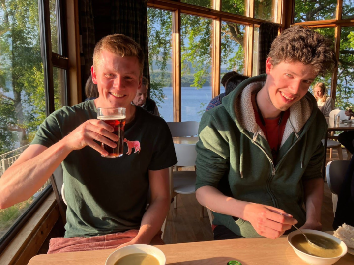 Two young men sit a table with beer. The one on the left has blond hair and the one on the right has brown, curly hair