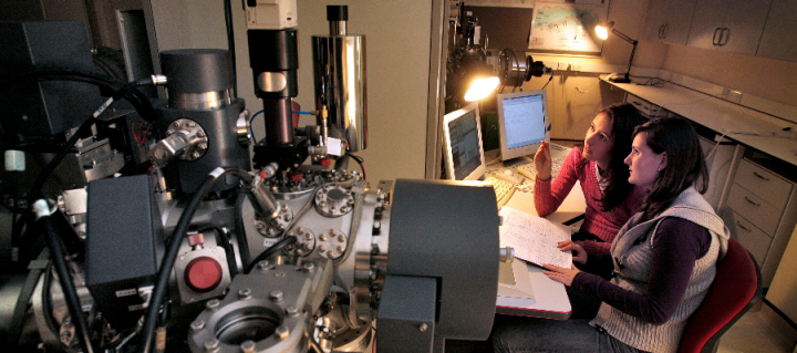 Two women seated at a desk looking at a computer and surrounded by technical equipment in an Earth sciences laboratory.