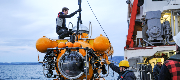 A male researcher in a wetsuit sitting atop a research sub being lowered from a ship into the sea
