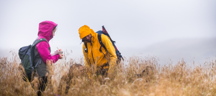 Two students in brightly coloured wet weather gear standing in long grass with grey rain and mist around them