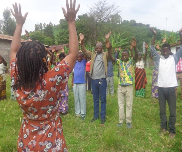 Photograph of a group of people doing capacitar practices in North Kivu, Congo. Their is a person with their back to the camera leading the practice, they are all standing with their arms stretched up over their head. 