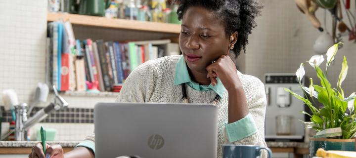 Student sat at her desk looking at a laptop and making notes in a notebook. 