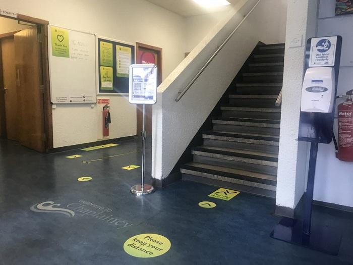 Photograph of the Chaplaincy foyer, there is green signage with the text "Please keep your distance". To the right hand side of the stairs is a hand sanitiser dispenser
