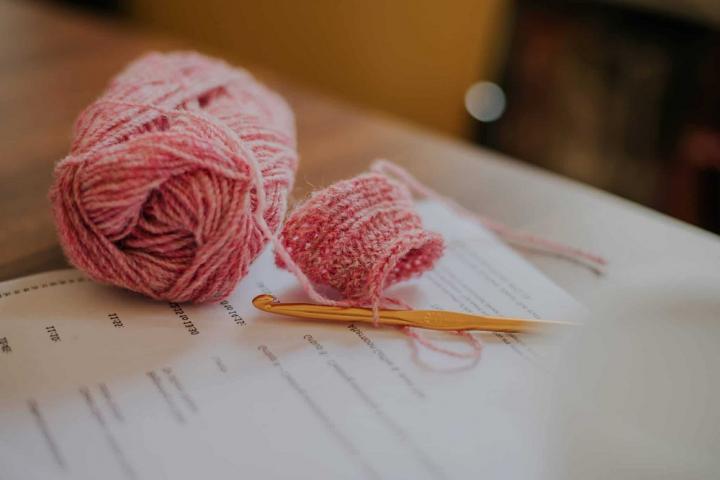 A ball of pink yarn and a small piece of fabric lay on a sheet of paper alongside a yellow crochet hook