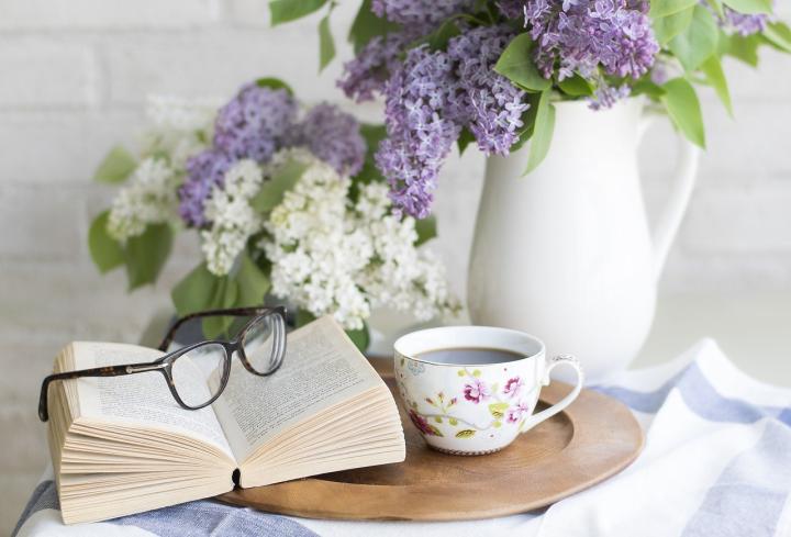 Photograph of an open book sitting on top of a wooden tray. On top of the book is a pair of brown reading glasses. Beside the book is a white cup with pink and green flowers on it, the cup is filled with coffee.
