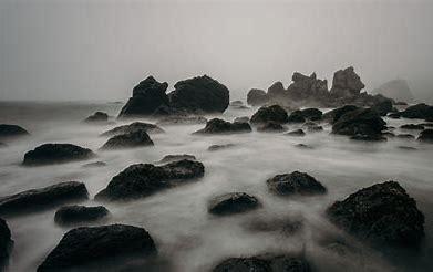 A black and white image of the mist rolling over the coastal rocks.