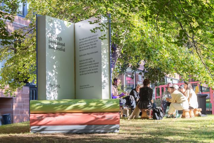 People sit on a bench outdoors next to a giant pile of books at Edinburgh International Book Festival