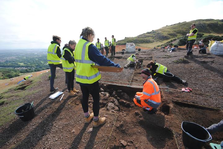 Students and professional archaeologists wearing high-vis clothing excavate and record a trench in Holyrood Park, Edinburgh