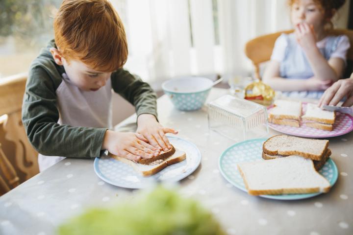 Young boy making their sandwich for lunch at a dining table as young girl watches in background