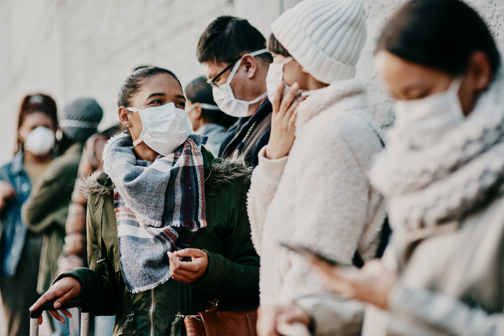 A group of young people standing interacting in groups while wearing protective face masks. 
