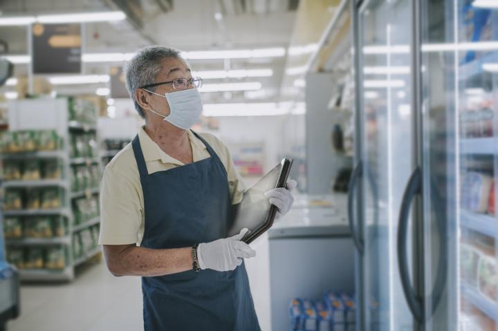 Supermarket worker who is wearing a mask and gloves and holding an ipad looks into a large food fridge.
