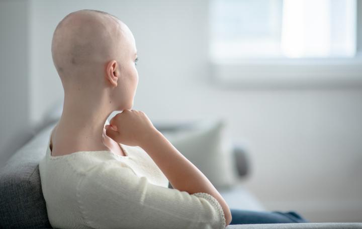 Young woman sitting on sofa, looking reflective. Her hair has been lost following chemotherapy.