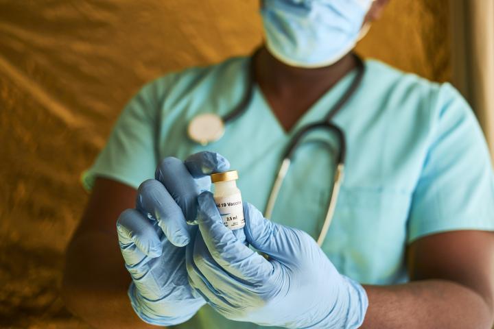 Image of African doctor holding a Covid-19 vaccine vial while wearing blue surgical gloves and scrubs