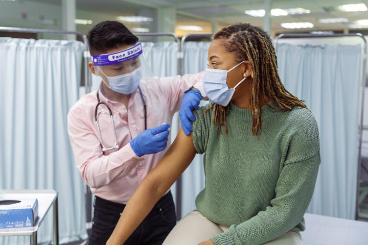 A young black woman wearing a protective face mask sits in a clinic as her male doctor of Asian descent prepares her vaccine