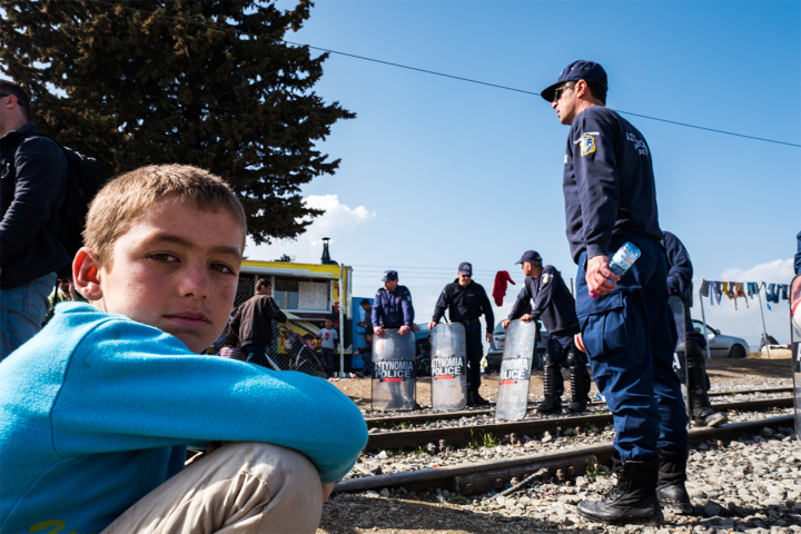Young boy sitting next to train tracks, surrounded by police men. He is wearing a blue t - shirt.