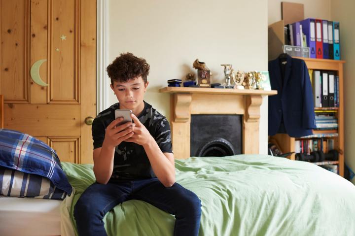 Teenage boy using smartphone, sat on the edge of his bed, with files and books on shelves in the background