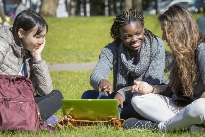 Three female students sit outside in the grass and work and talk around a laptop 