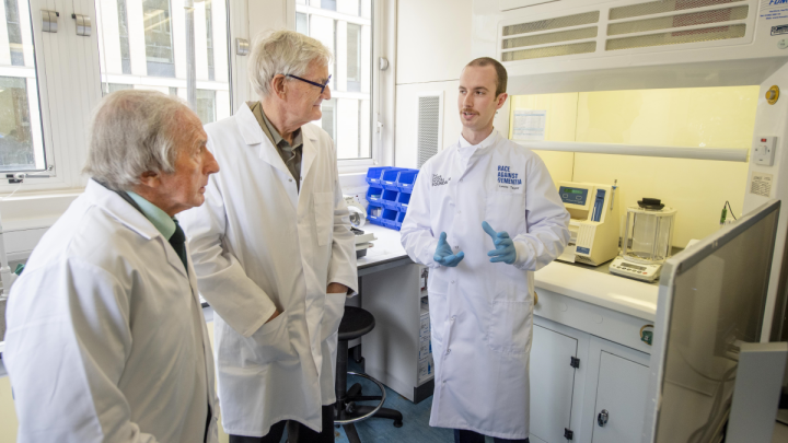 Sir Jackie Stewart and Sir James Dyson talking to Lewis Taylor in the lab wearing white lab coats