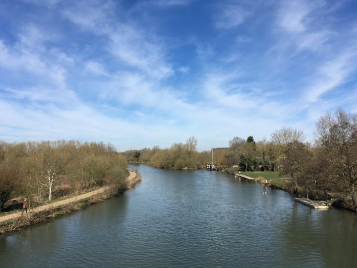 Photograph of the Thames in Oxford, on either side there is the banks of the river with bare trees and overhead there is blue skies.