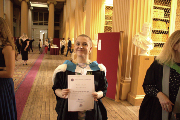 Emma Henriksen holds her award certificate up to the camera whilst smiling. She is wearing the Nursing Studies Graduation robes
