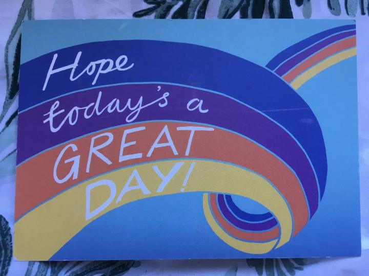 A photograph of a postcard. The background of the postcard is blue and there are different coloured strands looping across the postcard. The text of the purple, blue, yellow and orange strands reads 'Hope today's a great day'