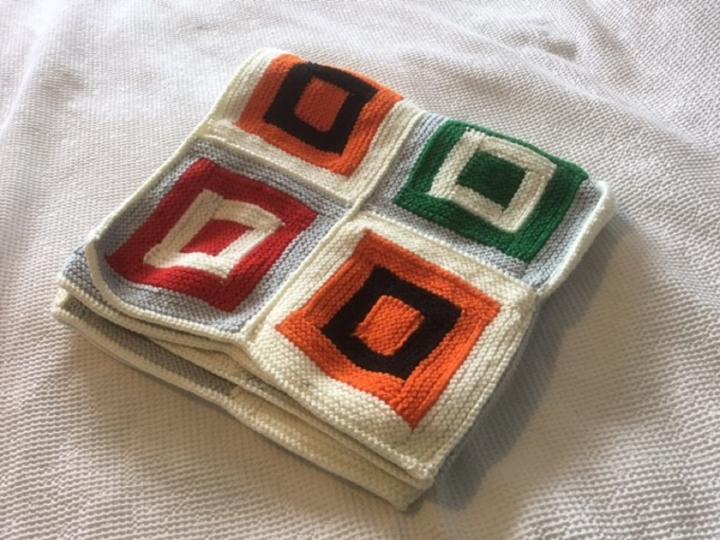Photograph of a knitted blanket lying on a white sheet. The blanket has 4 different coloured squares on it. 