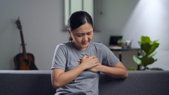 Asian woman using hands to clutch her chest in pain, sitting on sofa in living room at home