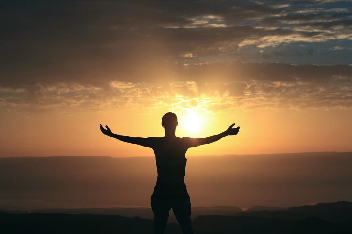 Photograph of a woman standing on a hillside admiring the sunrise in the distance. The silhouette is standing with her arms outstretched.