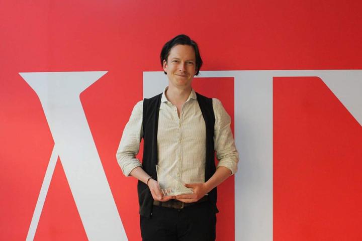 Photo of Patrick Errington holding his Emerging Writers award in front of a red and white screen