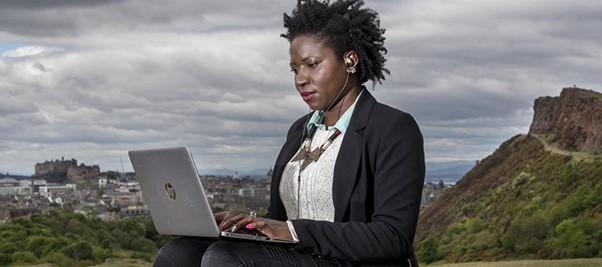 A woman sits in Holyrood Park, with Edinburgh Castle behind her to her left and Salisbury Crags her right, typing on a silver laptop which is placed on her lap.