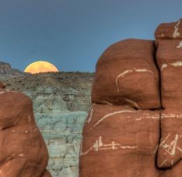 Photograph of brown rock formations in the foreground, in the background the moon is rising over the hills. 